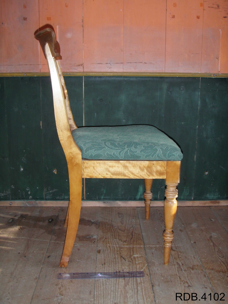 Stol i tre, lakkert. Fire føtter; framføttene er runde/dreiet, mens bakføttene er firkantede. Alle føtter er bredest øverst, og smalest nederst. Bakføttene fortsetter oppover, og er en del av stolryggen. Stolryggen har en støtte øverst, og en støtte midt på. Støtten øverst er firkantet. Støtten midt på har en runding midt på, og mot sidene går det to firkanter. 
Stolputa er løs, men sitter godt på fordi den er festet til noen spor på undersiden. Den er bredest frampå, og smalest bakerst. Stolputa er av tre, og trekt med et fôret, grønt stoff. 