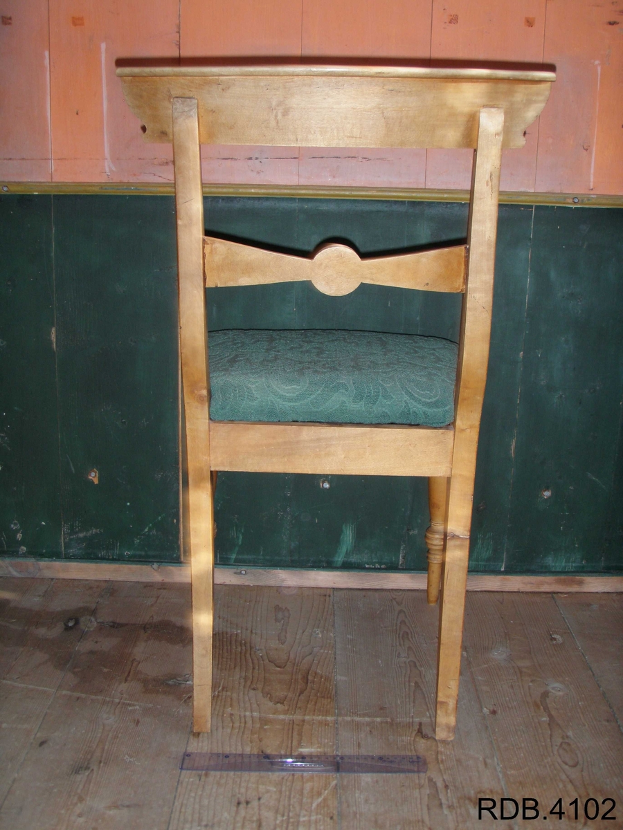 Stol i tre, lakkert. Fire føtter; framføttene er runde/dreiet, mens bakføttene er firkantede. Alle føtter er bredest øverst, og smalest nederst. Bakføttene fortsetter oppover, og er en del av stolryggen. Stolryggen har en støtte øverst, og en støtte midt på. Støtten øverst er firkantet. Støtten midt på har en runding midt på, og mot sidene går det to firkanter. 
Stolputa er løs, men sitter godt på fordi den er festet til noen spor på undersiden. Den er bredest frampå, og smalest bakerst. Stolputa er av tre, og trekt med et fôret, grønt stoff. 