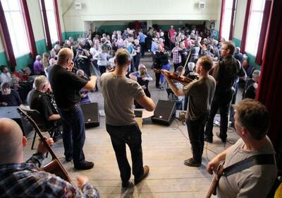 Her ser vi fra ei scene og ned på publikum. Vi står bak bandet, som spiller feler, gitar og bass. Nedenfor scena danser publikum.