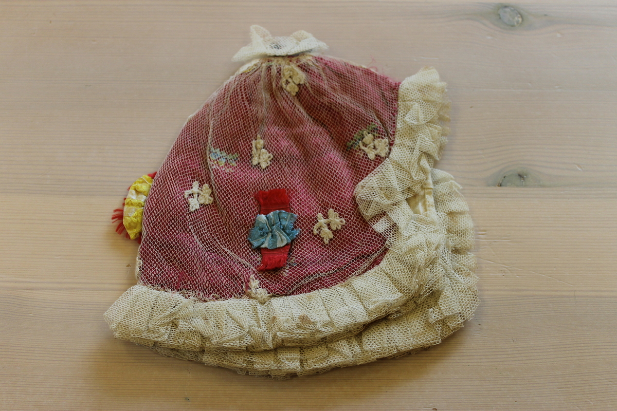 Rød dåpslue sydd av ett stykke, sammenrynket til en sirkel i nakken. Lua er trukket med hvit tyll, og har sammenrynket tyllkant rundt hele lua. Lua er pyntet med broderte blomster og sløyfer i flere farger. Fôret med hvitt bomullsstoff. 