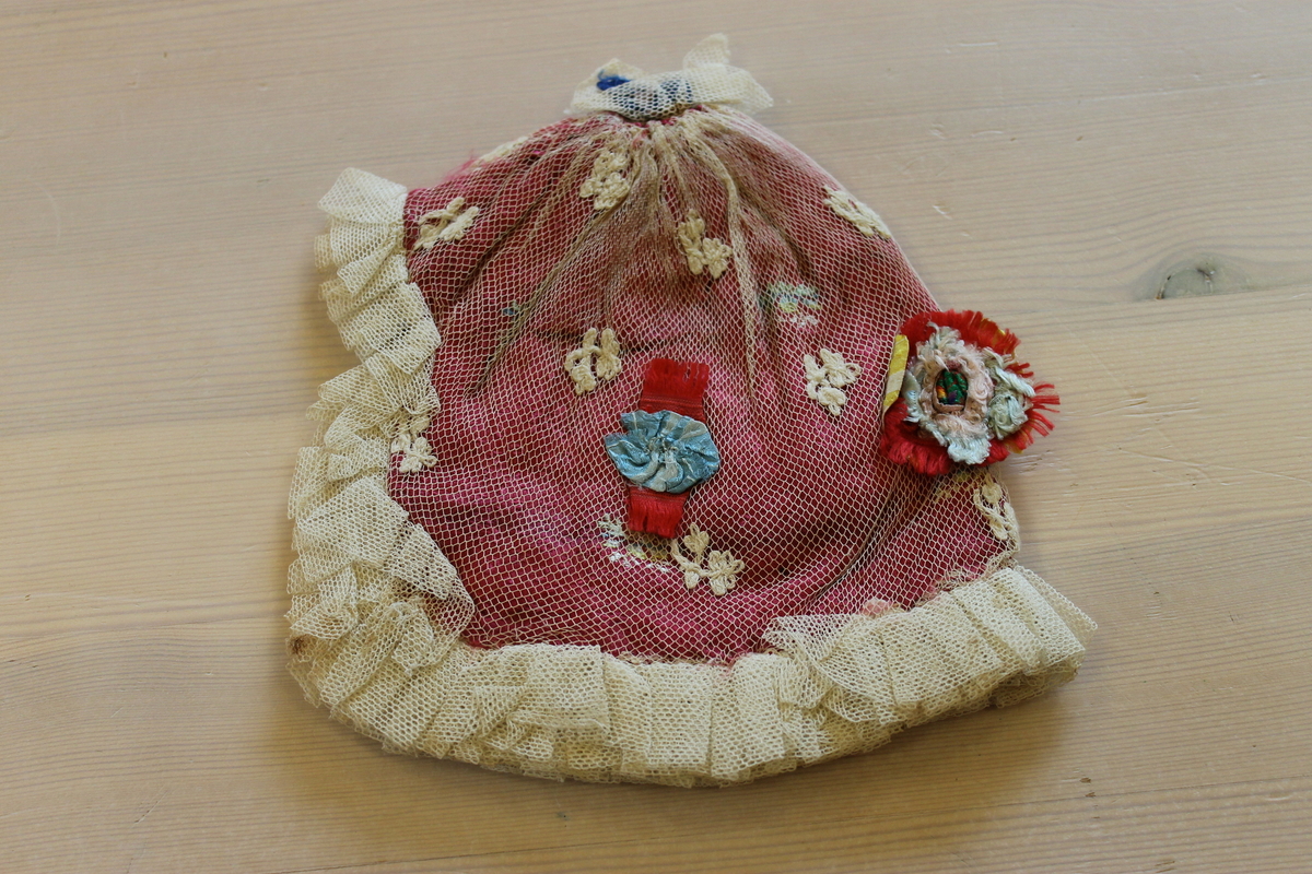 Rød dåpslue sydd av ett stykke, sammenrynket til en sirkel i nakken. Lua er trukket med hvit tyll, og har sammenrynket tyllkant rundt hele lua. Lua er pyntet med broderte blomster og sløyfer i flere farger. Fôret med hvitt bomullsstoff. 