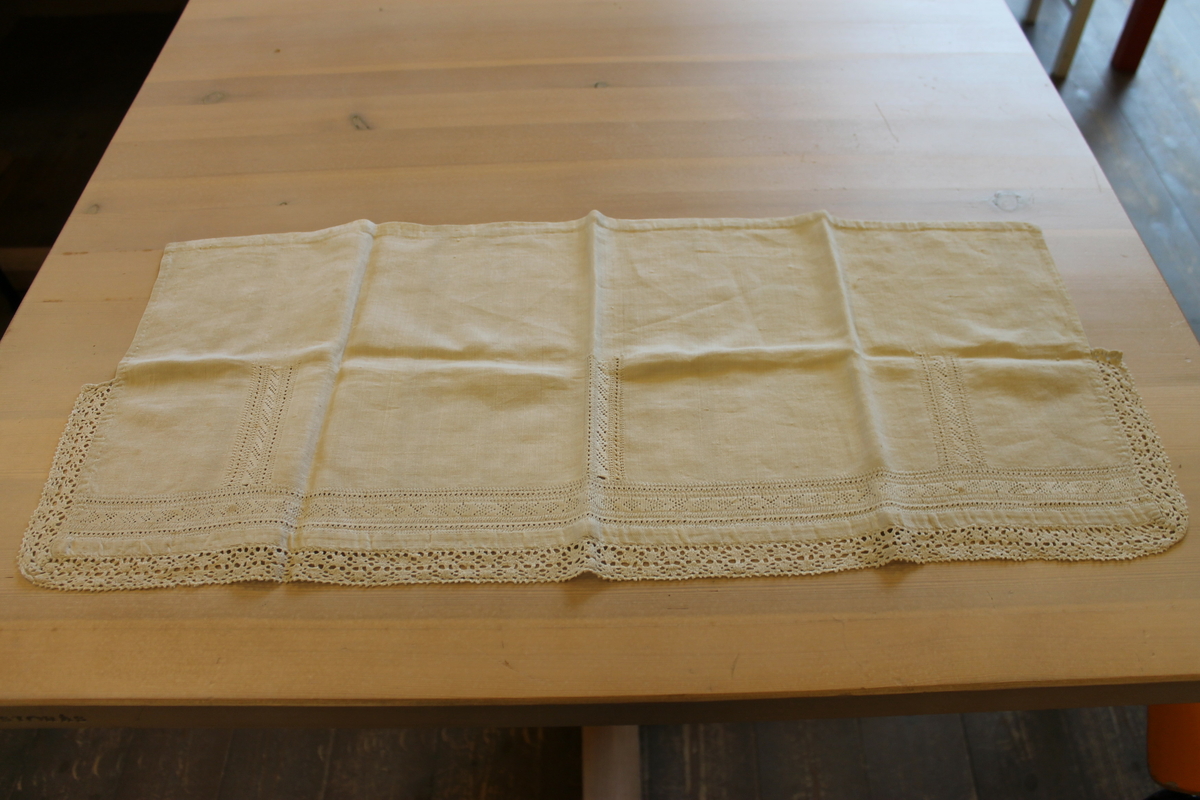 Rektangulært stykke lin (mål: 30,5 cm x 70cm ). Den ene halvdelen (brettet på langs) er rikt dekorert med tellesøm og kantet med bomullsblonde (trolig ikke håndlaget), mens den andre halvdelen, som blir brettet inn, er uten dekor