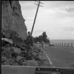 Rasberget vid länsgränsen 1961. En man står med ett spett vid en bergvägg. Runt honom finns rasmassor. Bakom honom står en elstolpe på sned. Bilar åker på vägen bredvid.