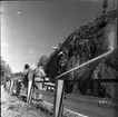 Rasberget vid länsgränsen 1961. En vattenkanon åker längst med en motorväg och har kanonen riktad mot en bergvägg. Vattnet träffar högt upp på berget och nedanför ligger rasmassor. Mot vägstaketet står tre herrar lutade, en av dem är polis.