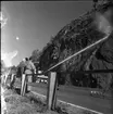 Rasberget vid länsgränsen 1961. En vattenkanon åker längst med en motorväg och har kanonen riktad mot en bergvägg. Vattnet träffar högt upp på berget och nedanför ligger rasmassor. Mot vägstaketet står tre herrar lutade, en av dem är polis.