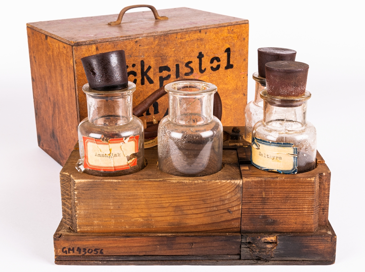 Låda med glasflaskor under masonithuv.
Tillverkare: Tegellaboratoriet vid ___ IVA
Glasflaskorna har innehållit bland annat Ammoniak och Saltsyra.