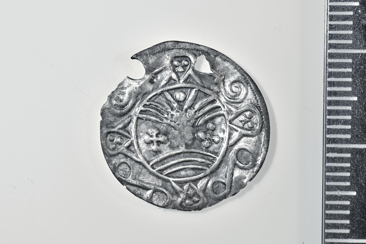 Motiv advers: Ansikt med stråler og bart. Fire mindre masker, to 8-tallsymbol og to slanger i ring rundt. 

Motiv revers: Hjort som vender framover, slange foran mulen, solsymbol bak og trikvetra symbol under. 