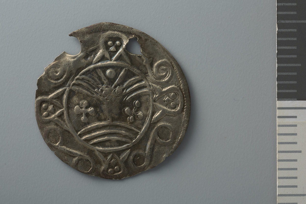 Motiv advers: Ansikt med stråler og bart. Fire mindre masker, to 8-tallsymbol og to slanger i ring rundt. 

Motiv revers: Hjort som vender framover, slange foran mulen, solsymbol bak og trikvetra symbol under. 