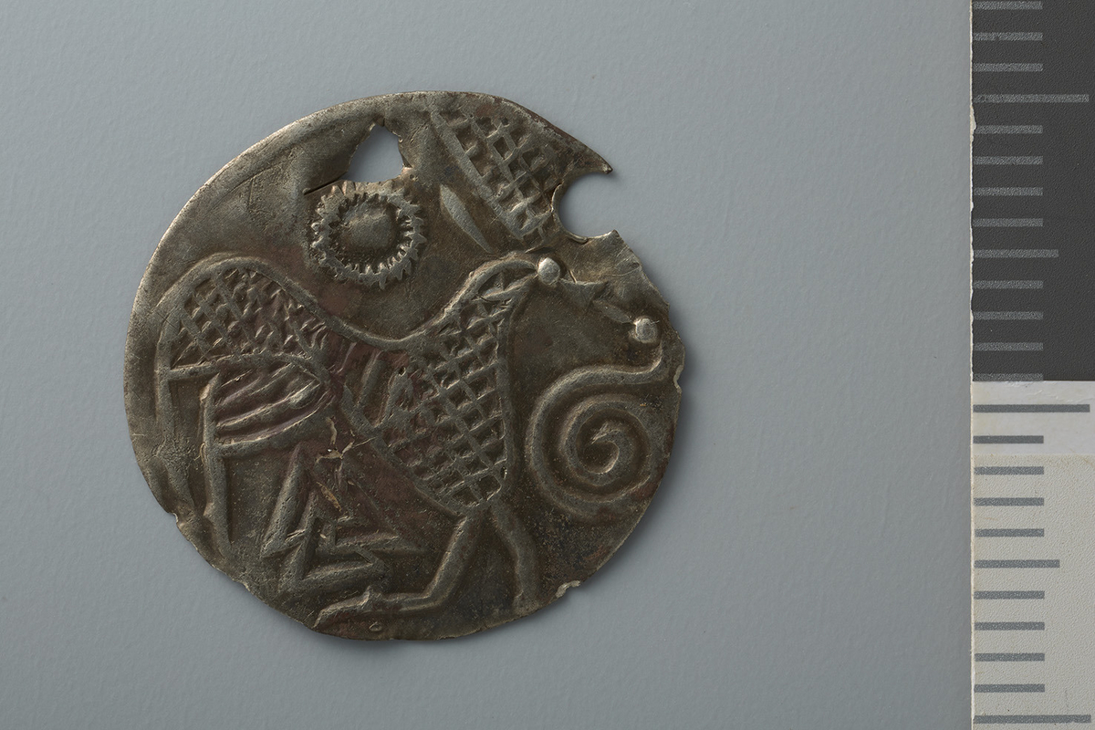 Motiv advers: Ansikt med stråler og bart. Fire mindre masker, to 8-tallsymbol og to slanger i ring rundt. 

Motiv revers: Hjort som vender framover, slange foran mulen, solsymbol bak og trikvetra symbol under. 