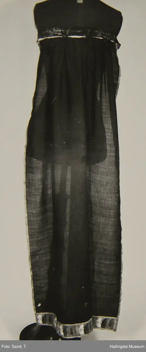 Fyriklæ i bomull med fløyelsband og ullband. Svart, tunt tøy i fyriklæet, liknar noko på bomull, men er noko stivare. Toskaft-veving. Rynka øvst, kvit bomullslist sett på øvst, br. 2,4 cm. Over denne festa ei bolu (svart fløyelsband med blomemønster i fiolett), l.: 38 cm og br.: 4 cm. Nedst eit tunt ullband, br.: 5,8 cm, blomemønster i 3 blåfargar, 3 grønfargar, kvitt og gult på raud botn. Langs sidene raudt ullband, 0,7 cm. Fyriklæet er sundt og bøtt sumstand.