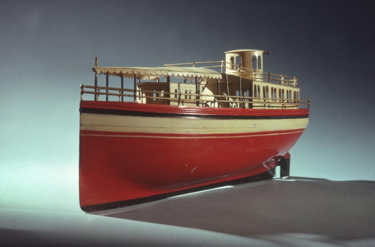 Fartygsmodell. Leksaksbåt av plåt. Rödmålat skrov med svartvitt bälte över vattenlinjen. Rak förstäv, häckakter, roder och en trebladig propeller. Förligt däckshus överspänt med "suntält" av plåt, akterut passagerarsalong, navigationshytt. Propellermaskineriet består av en tvåpolig elektrisk motor driven av ett De Lanche element. Gråmålat skrå.