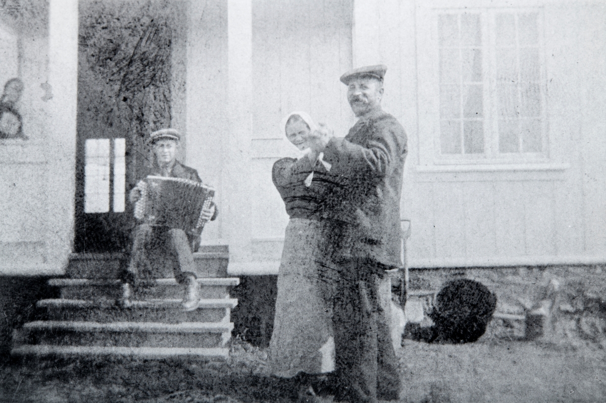 Spillemann Eivinf Hagen (1901-1990), spiller på trekkspill. Mens Nils Hagen (1862-1955) og Eline Hagen (1857-1941) tar seg en dans på tunet ved husmannsplassen Hagen, under Hverven Gård i Ottestad.