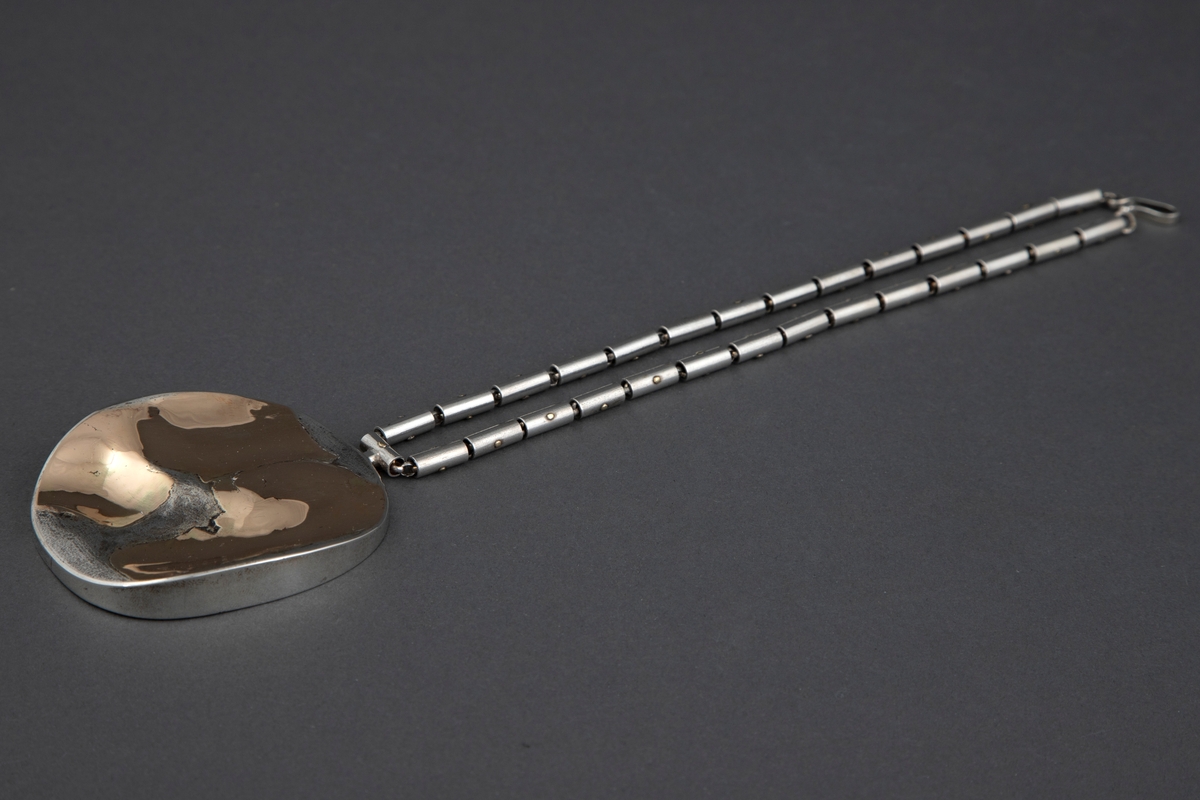 Anheng i sølv i tilnærmet elipseform med påsmeltede gullplater. Smykket har en buklet overflate der blankt gull kontrasterer med matt sølv. Kjede av små pånittede rørstykker av sølv.