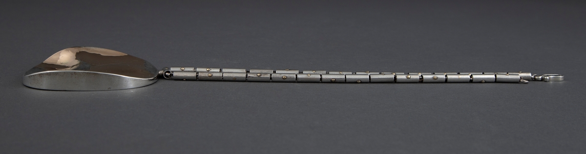 Anheng i sølv i tilnærmet elipseform med påsmeltede gullplater. Smykket har en buklet overflate der blankt gull kontrasterer med matt sølv. Kjede av små pånittede rørstykker av sølv.