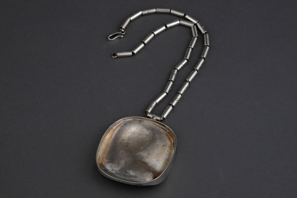 Anheng i sølv i tilnærmet elipseform med påsmeltede gullplater. Smykket har en buklet overflate der blankt gull kontrasterer med matt sølv. Kjede av små pånittede rørstykker av sølv.