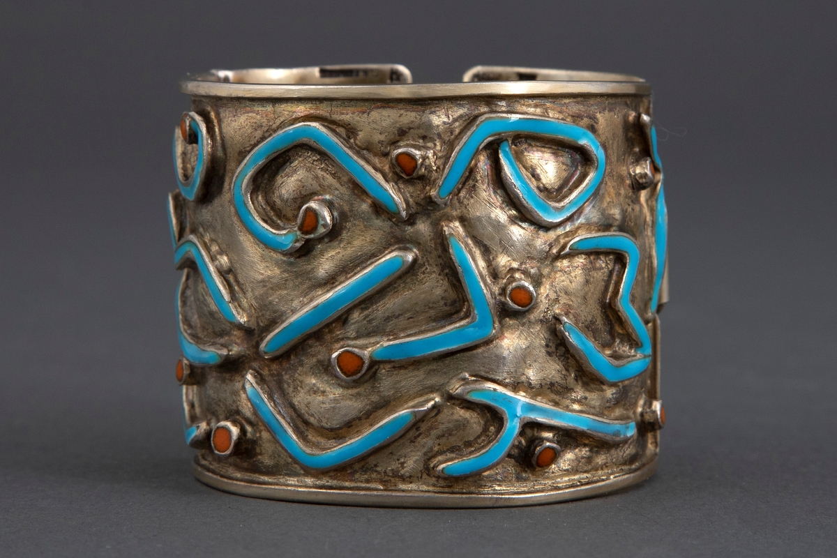 Bredt armbånd i forgylt sølv med emalje. Ujevn overflate med opphøyede, uregelmessige båndornamenter dekket med turkis emalje. Innimellom er det små opphøyede punkter med brunoransje emalje. Armbåndet har lås på siden.