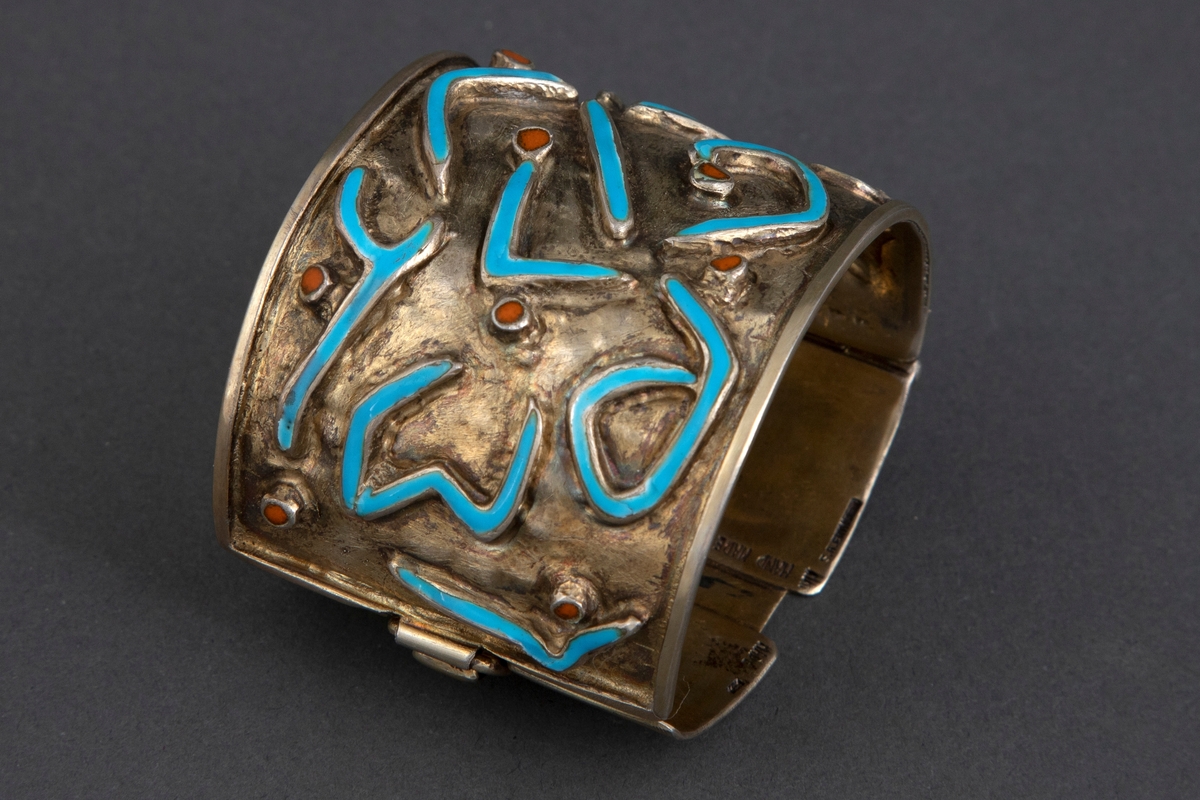 Bredt armbånd i forgylt sølv med emalje. Ujevn overflate med opphøyede, uregelmessige båndornamenter dekket med turkis emalje. Innimellom er det små opphøyede punkter med brunoransje emalje. Armbåndet har lås på siden.