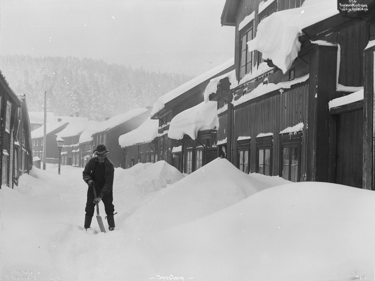 Mann måker snødekkt gate i Trangvik, snøfonner opp mot husvegg. Fotografert januar 1902.