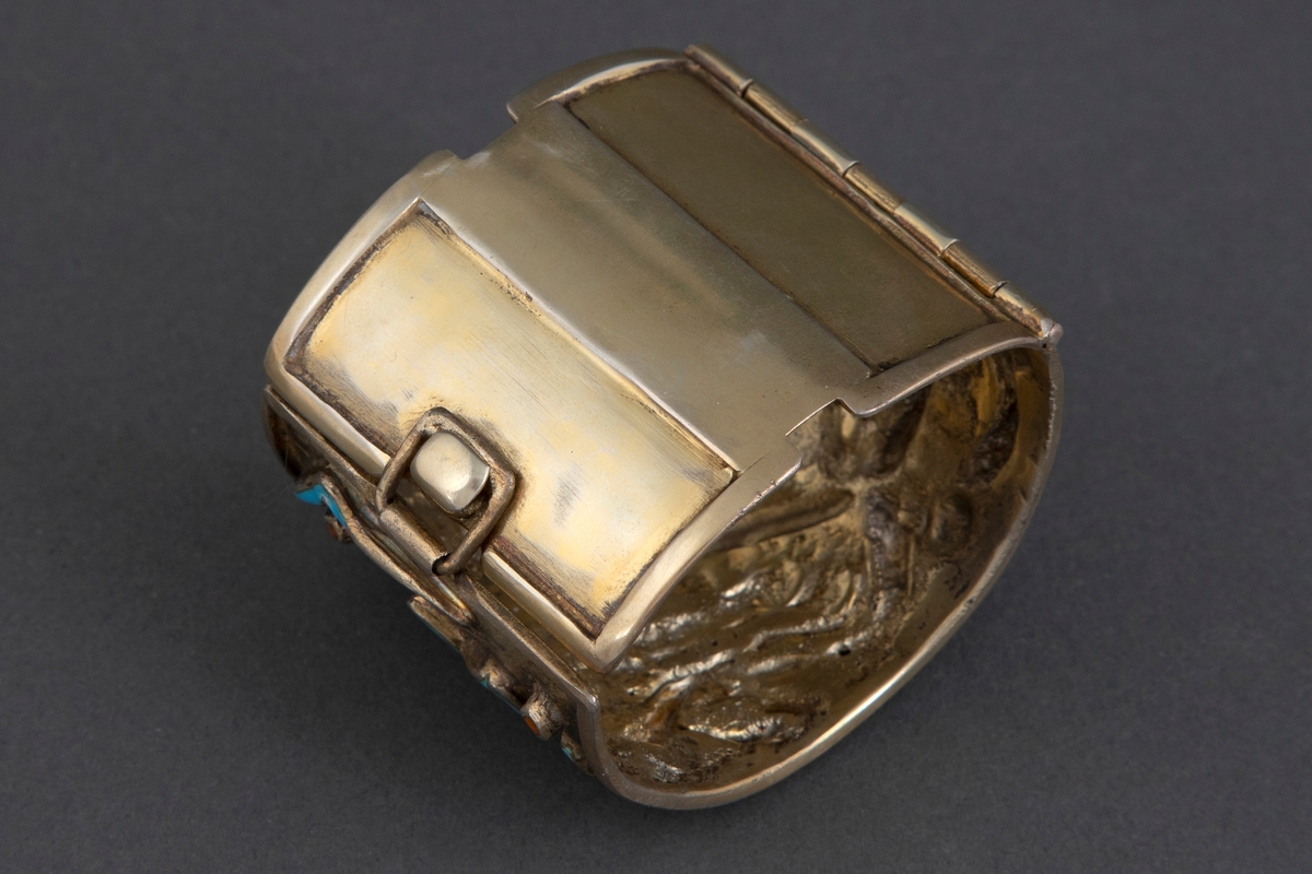 Bredt armbånd i forgylt sølv med emalje. Ujevn overflate med opphøyede, uregelmessige båndornamenter dekket med turkis emalje. Innimellom er det små opphøyede punkter med brunoransje emalje. Armbåndet har lås på siden.