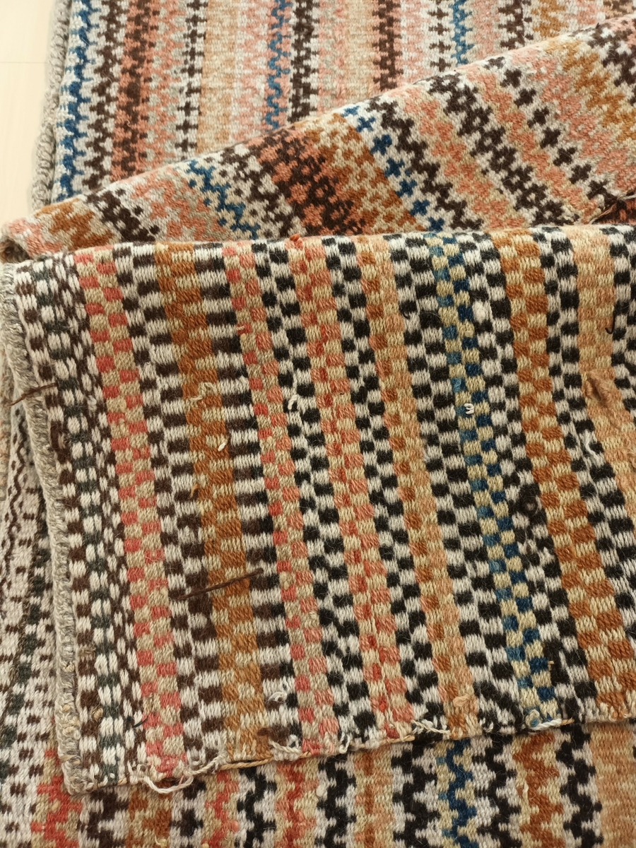 Teppe i krokbragd i mange fargar. Renning: 26/10 cm. tvunnet lintråd. Innslag: 2trds ullgarn. Teppet er skjøtt på midten, og bordane møtes tilfeldig i skøyten.