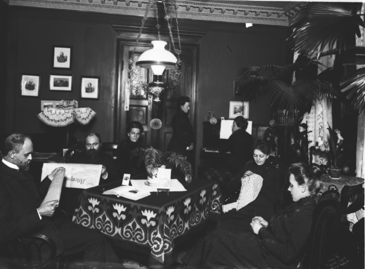 Familien samlet i stuen hos Wilse, diverse hjemlige sysler som brodering, pianospill, avislesning. Fotografert 1902
.