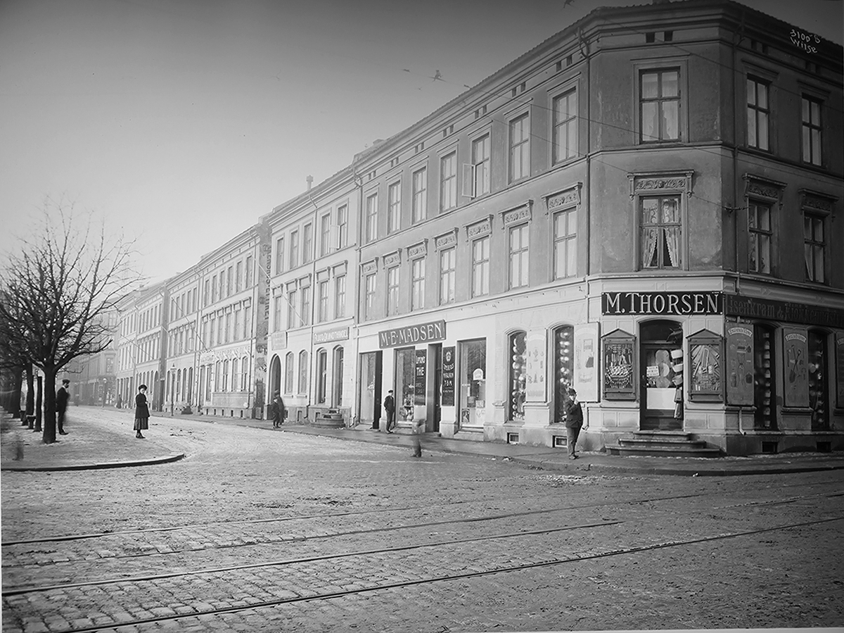 Del av husrekke Olaf Ryes plass. Butikk på hjørnet, i forgrunnen gate med trikkeskinner.