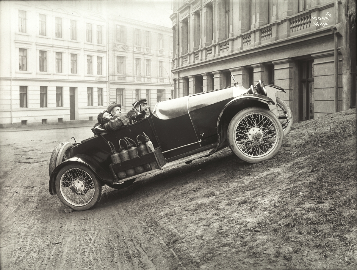 Tre menn i sportsbil, alle tidsmessig antrukket.  Bilen er parkert i et bymessig område.