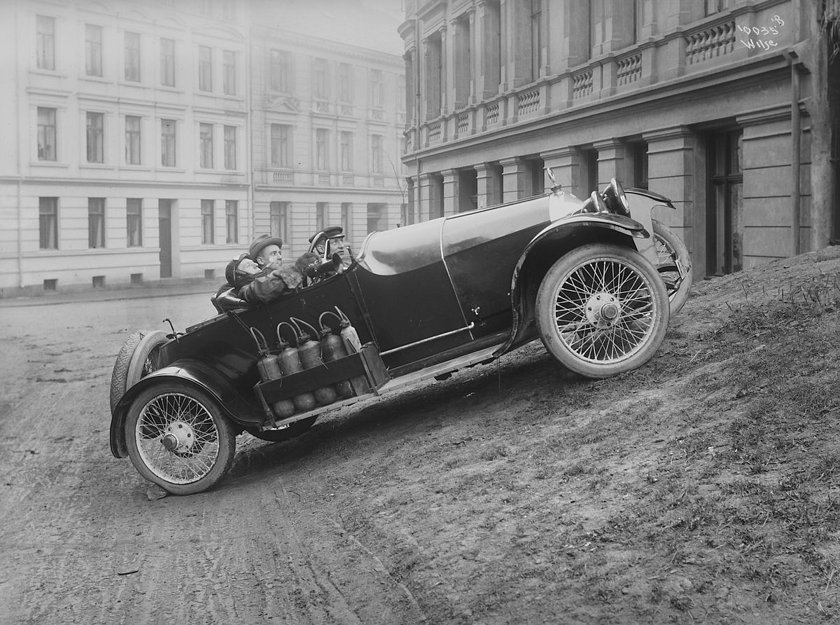 Tre menn i sportsbil, alle tidsmessig antrukket.  Bilen er parkert i et bymessig område.