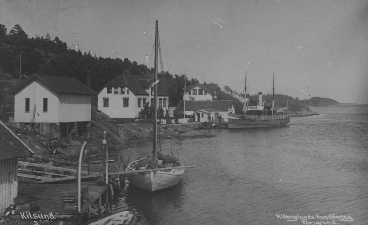 Hus ligger langs fjorden med ferge og seilbåt ved brygge, Kilsund. Påskrevet "H. Wergelands Kunstforlag, Porsgrund", "Kilsund". Fotografert 1922.