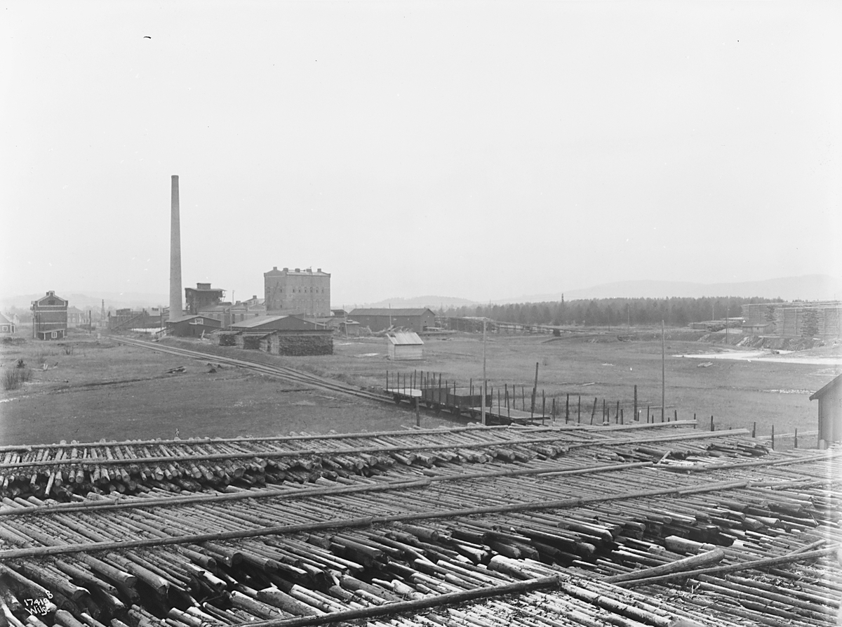 Masse tømmer ligger lagret foran fabrikkområde, Lillestrøm cellulosefabrikk. Fotografert 1923.