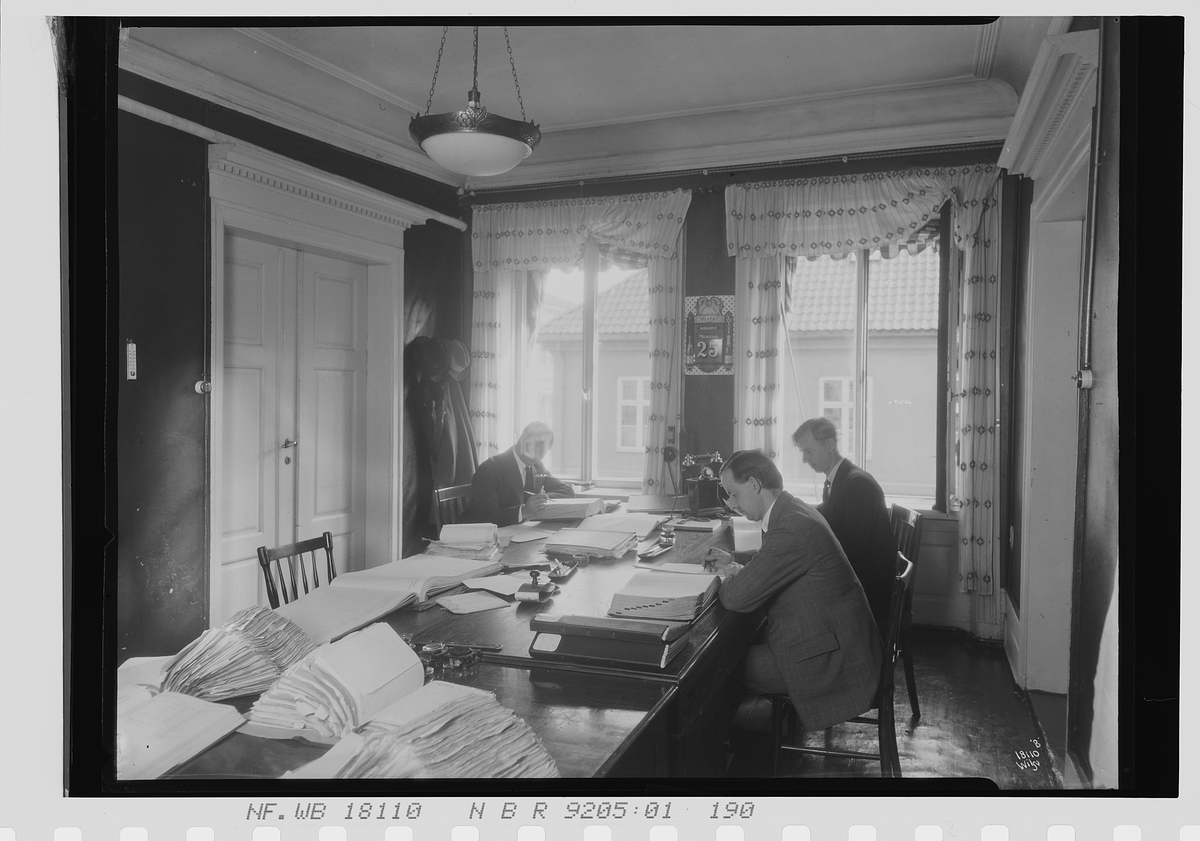 Menn sitter ved stort bord med oppslåtte bøker, Vinmomopolet. Fotografert 1924.