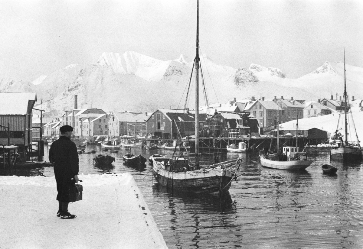 Mann med dokumentmappe står på bryggen, litt usikkert om han er på skyss eller om han venter tilreisende.  Fjell i bakgrunnen og båttrafikk langs bryggekanten.