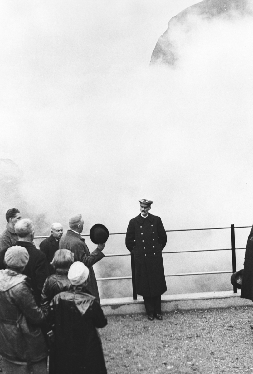 Kong Haakon med følge vged gjerdet som omgir utsiktspunktet med Trollstigen og omgivelsene.