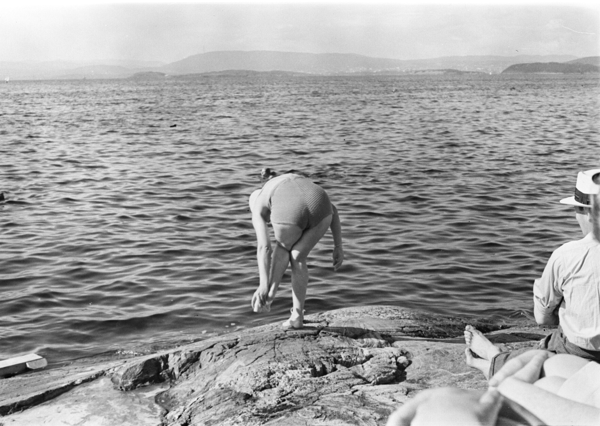 Svaberg og sjø, kvinne i ferd med å ta av skoene og hoppe i vannet. Mann med stråhatt nyter sommer og sol.