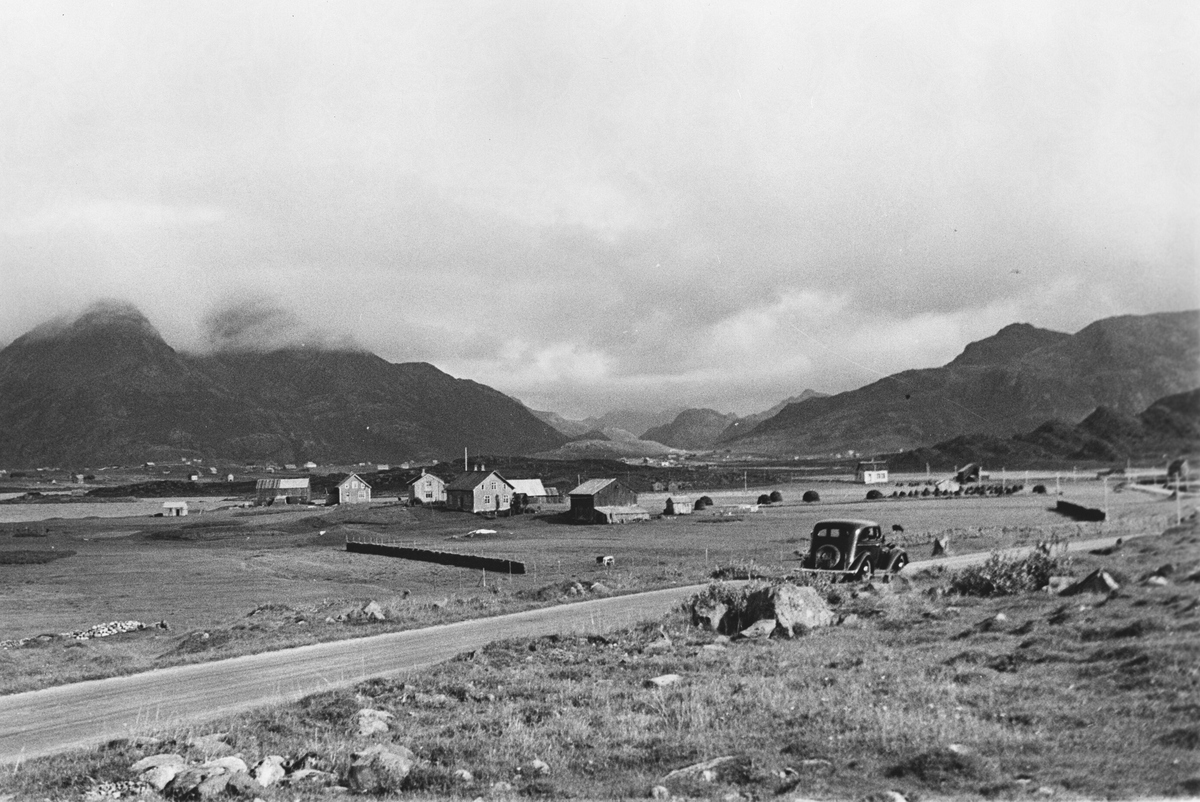 Kystlandskap med bebyggelse langs veien, fjell i bakgrunnen, personbil på veien.