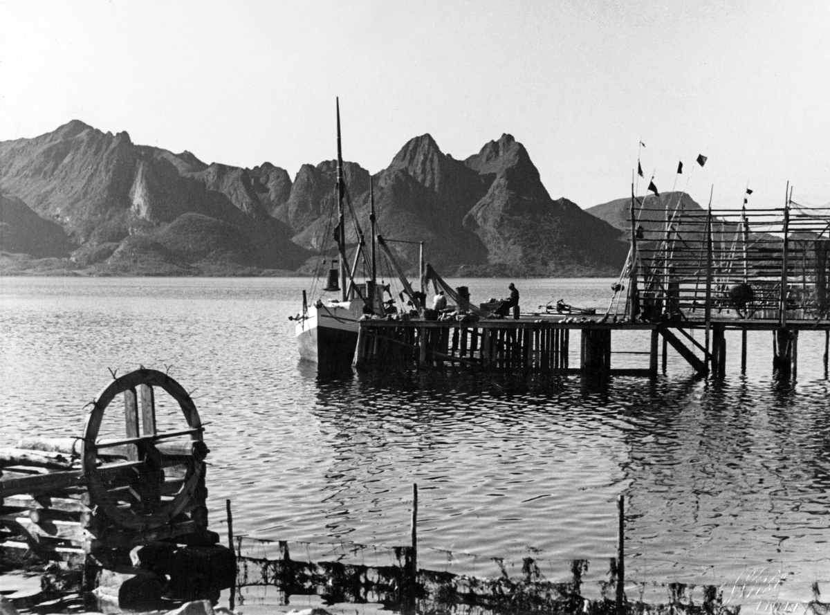 Fiskebåter ligger til ankers langs bryggene, I bakgrunnen høye fjell langs fjorden.