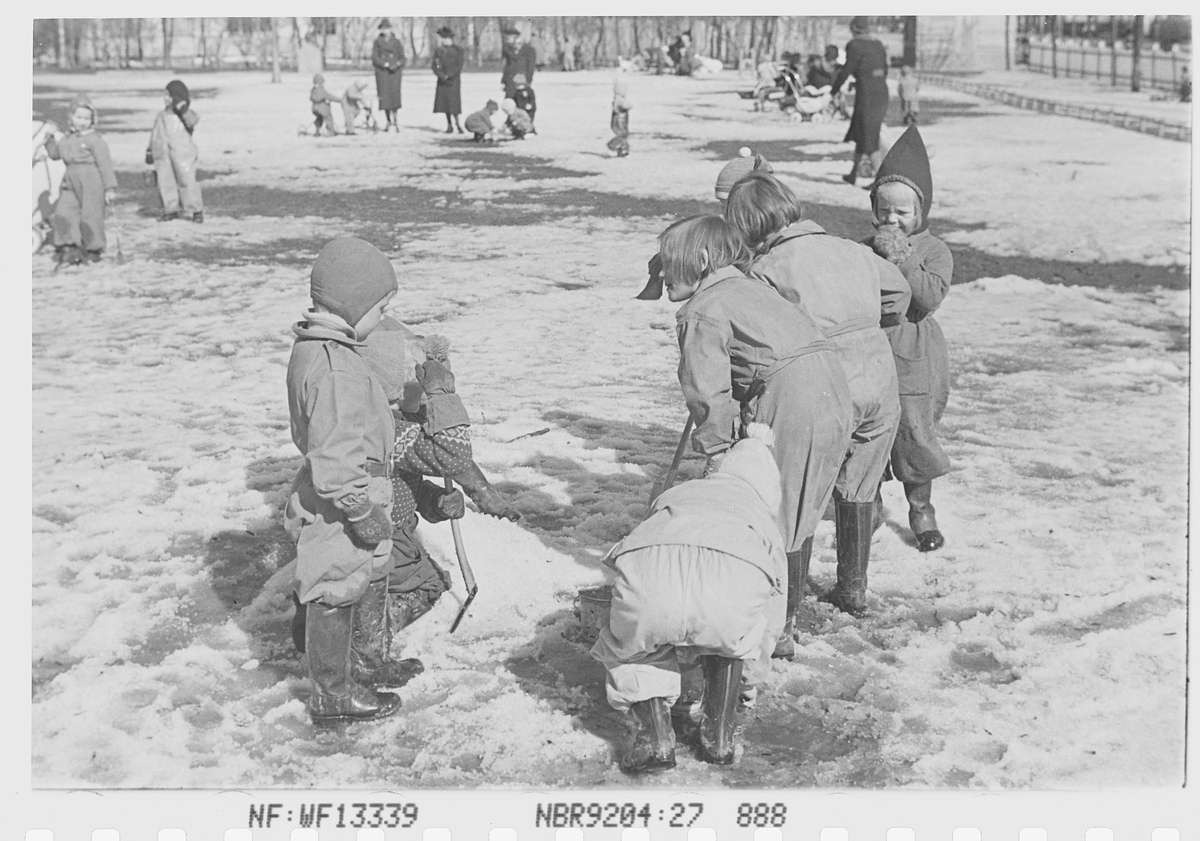 Barn leker med spader i snøen på lekeplass. Fotografert 1940.