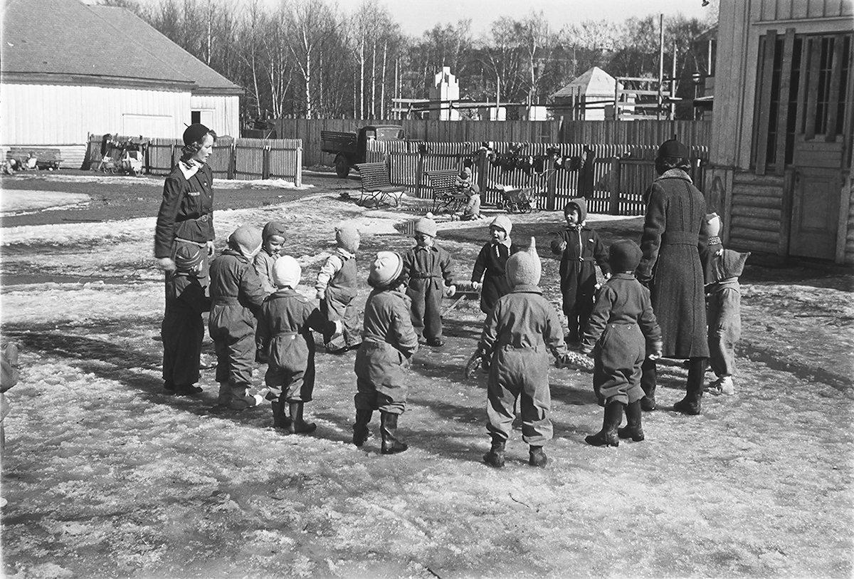 To voksene damer med barnegruppe som står i ring på lekeplass. Fotografert 1940.