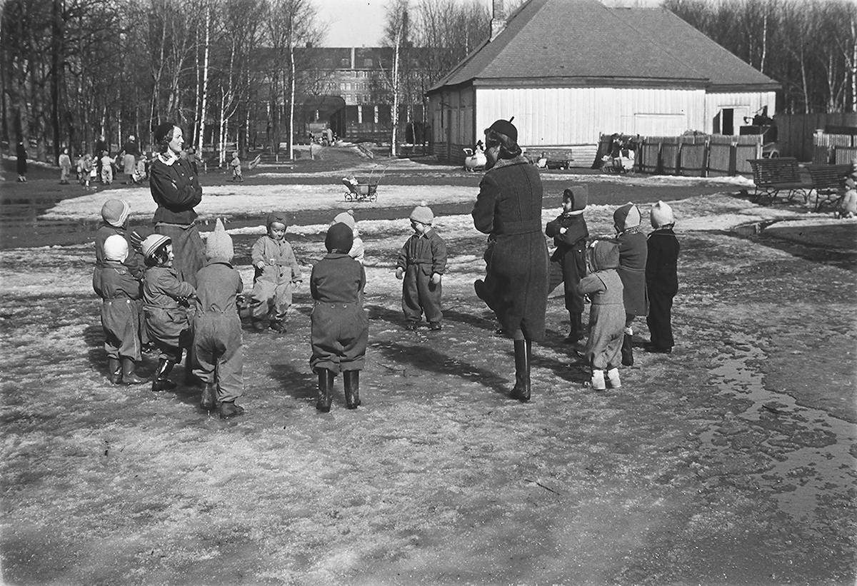 To voksene damer med barnegruppe som står i ring på lekeplass. Fotografert 1940.