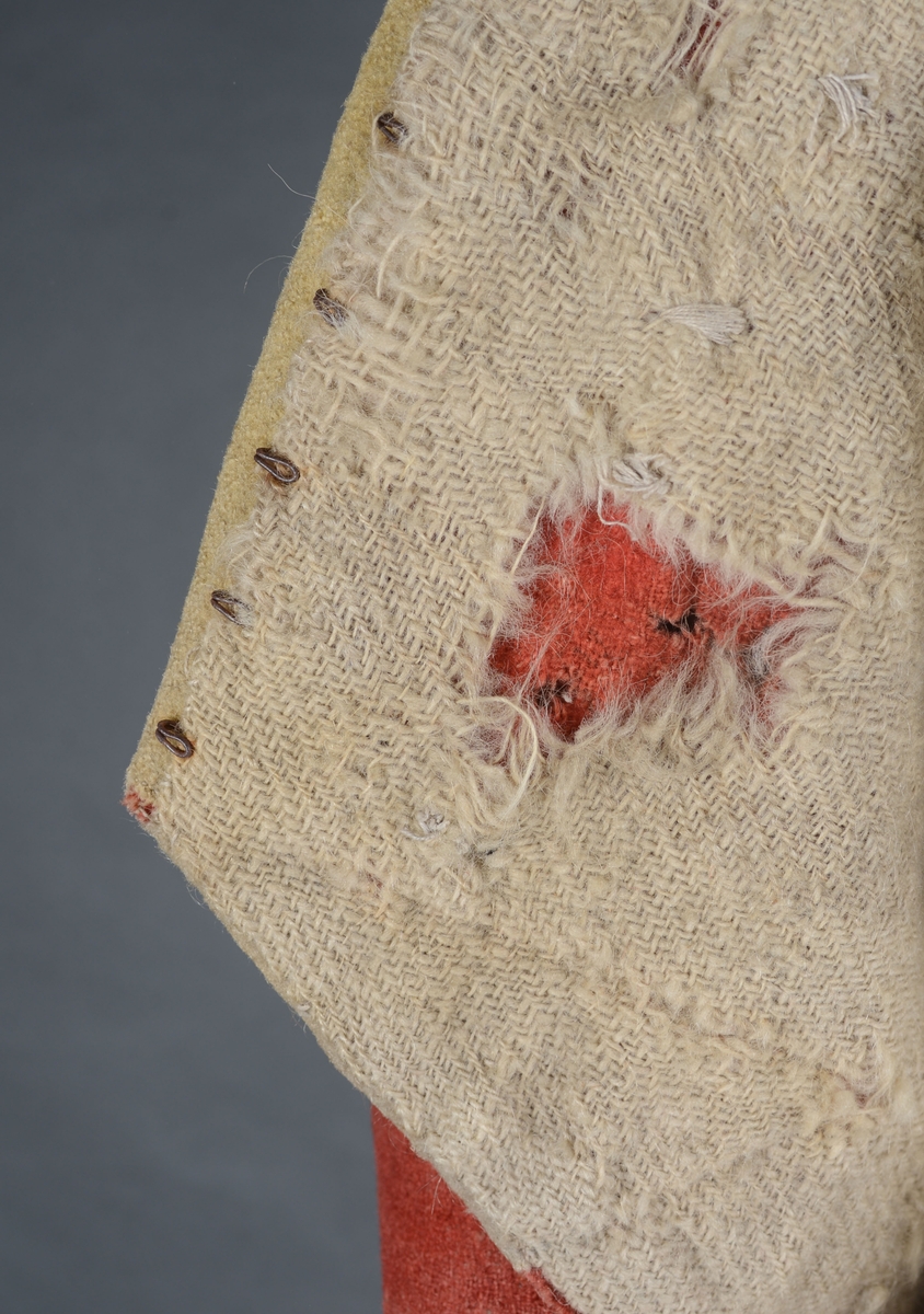 Kort trøye med høy ståkrage, sydd i grovt rødt lett valket ullstoff i empiresnitt. Trøya er helforet med grovt ullstoff. Kragen har mellomfor som er sydd fast til innerkragen. Ytterstoffet på kragen er brettet over på innsiden i øvre kant. Tittekant med rødt stoff i kortsidene på kragen. Trøya har søm MB og bakovertrekte skulder- og sidesømmer. Sidesømmene buer fra ermrigningen og mot MB. Sidesømmene slutter som legg med innlegg av gult stoff. Samme stoff brukes også som ytterkrage og slag på framstykket. Slagene er fastsydd og går opp som trekantforer mot kragen. Kneppes fast i bolen. 7 knapphull, men det mangler mange knapper. Plagget lukkes med 8 par metallhekter MF.  Nedenfor slagene er det påsydd trekanter av gråbeige ullstoff. Fra slagene foran skrår trøya bakover. Tosømsermer med dekor frampå ermene. Påsydd gult stoff med 2 striper av lys gråhvite bånd som går opp i en spiss mot innersømmen på ermet. Splitt ved yttersømmen. Denne lukkes med trekt rød knapp og knapphull. Mangler knappen på venstre erme. Stikninger langs slagene og kragen. Trøya er helt håndsydd og ytterkantene er sydd med sårkant på både for og stoff Tekst på 1 knapp, 3 knapper med stripe effekt inn mot midten.
Rød stoffhempe i nakken MB. Trøya har trolig tilhørt militæruniform fra tidlig 1800tall.