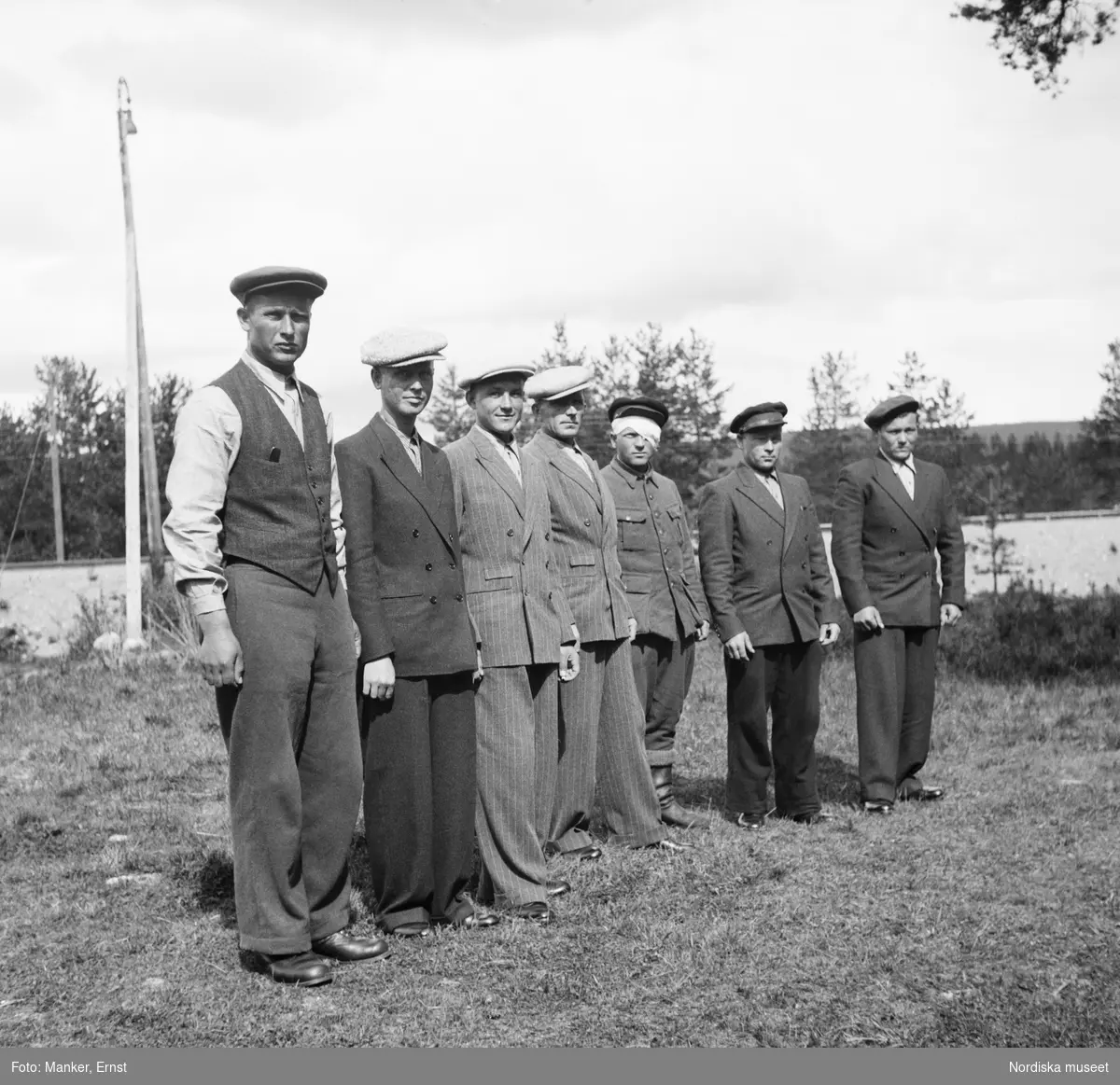 En grupp män står på rad framför kameran. Sju ryska flyktingar kom med buss från Björkholmen till Jokkmokk i sällskap med Ernst Manker. De har just besökt Josefssons klädaffär och ekiperat sig inför avfärden till interneringslägret i Huddinge. Som flyktingar i Sverige fick de inte använda sina militära uniformer. Från höger till vänster Ivan Pakatilov, Gregority Babykin, Jakov Olejnik, Ivan Patapov, Aleksej Martjenko, Nikolaj Tsopkalo och Gregorij Vachotin.