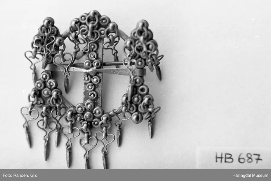 Byggkornsølje, sølv. Ring med tverribbe og 2 nåler. Øverst og nederst på ribben samt over og under begge nåler rombeformet plate dekket av 9 kuler med riftlet rand, hver rombe har 3 byggkorn, dobber. De 2 nåler danner tilsammen en rett linje. Dobbene består av en herteformet bøyle med en løkke nederst hvori der henger en liten tapp.
R.B.S.