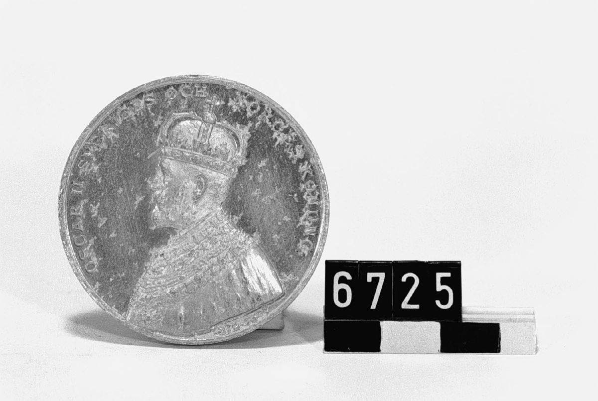 I aluminium. Tjocklek: 4 mm. Åtsidan: Vänster profil av Oscar II och "Oscar II Sveriges och Norges konung". I nedre kanten "1872-1897". Frånsidan: Bild av utställningshall samt text runtom: "Minne af Allmänna Konst- och Industriutställningen i Stockholm 1897". Signerad Lea Ahlborn. Angripen av aluminiumpest.