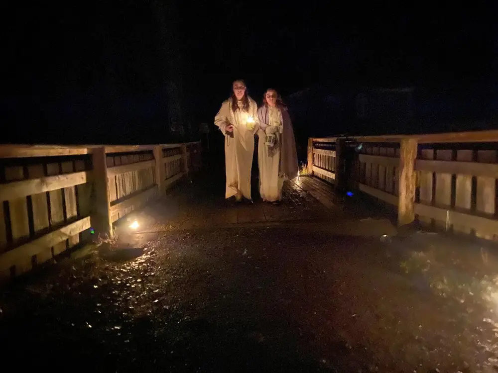 To hvitkledde skikkelser med en lykt på vei over ei bru. Det er helt mørkt rundt dem. Skummel stemning.