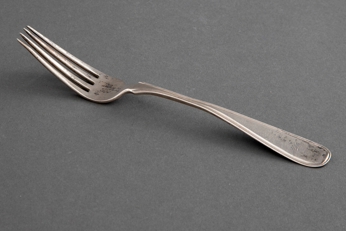 Gaffel i sølv. Jevnt utvidet skaft, kun dekorert med ei smal kantstripe. Med etui, hvor tilhørende gaffel også finnes.