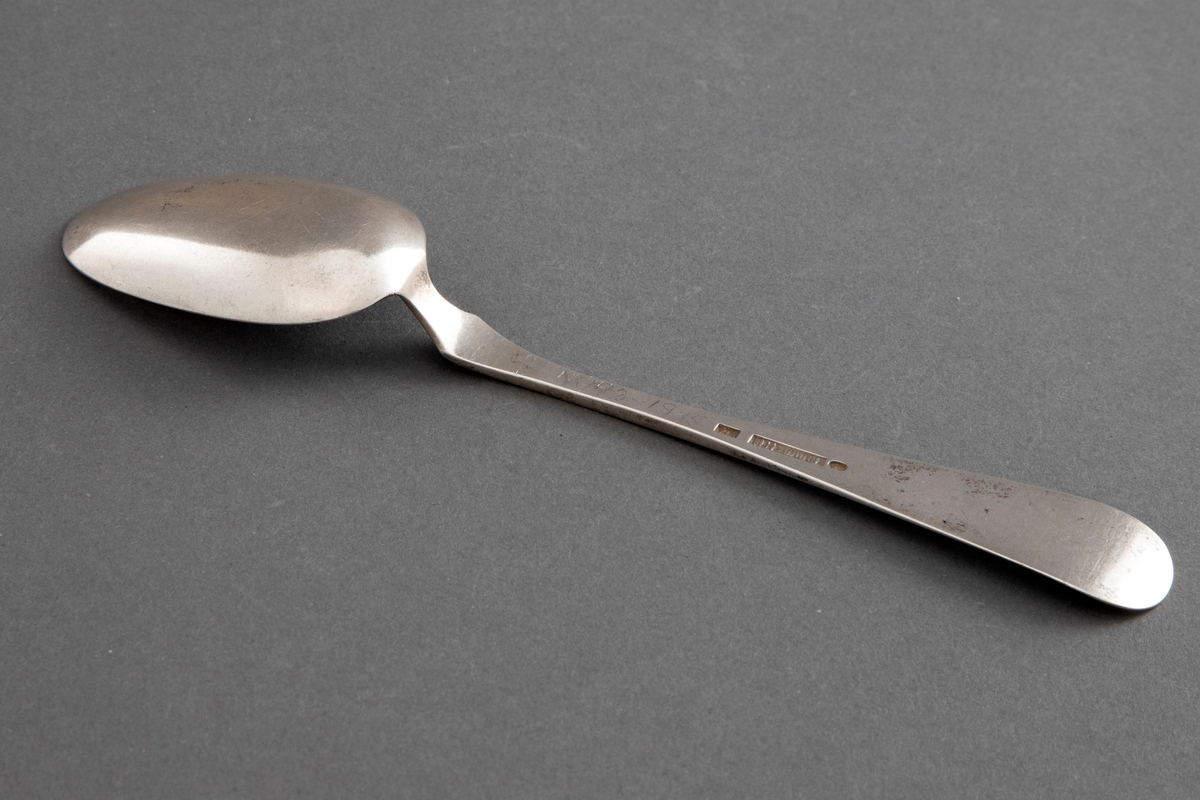 Skje i sølv. Jevnt utvidet skaft, kun dekorert med ei smal kantstripe. Med etui, hvor tilhørende gaffel også finnes.