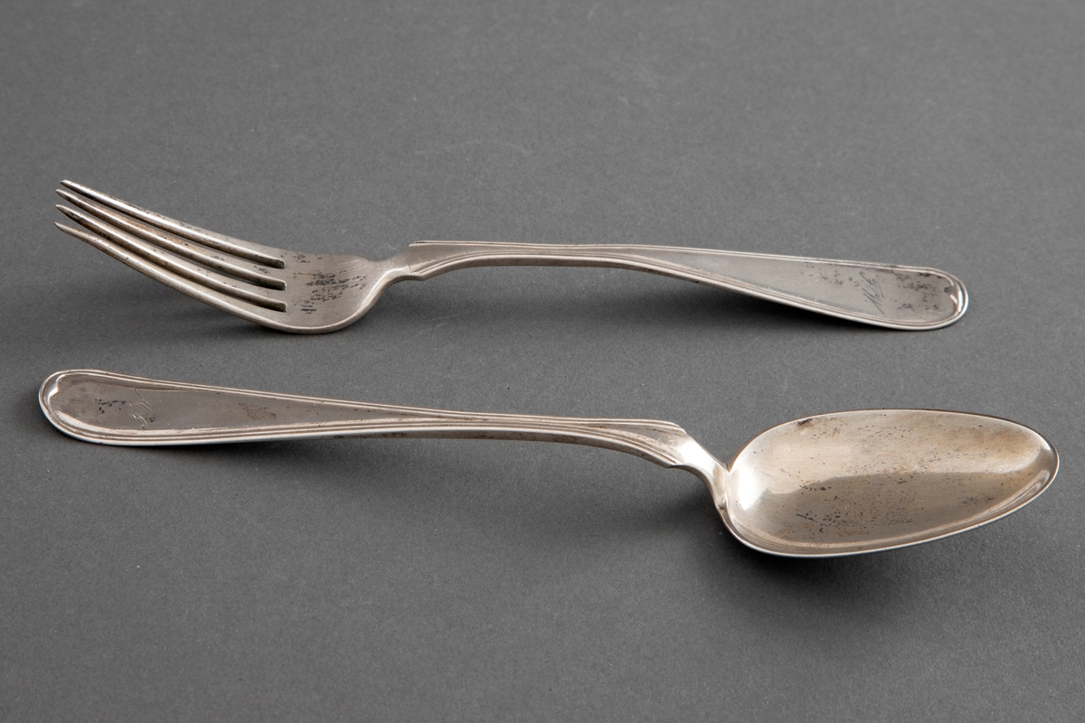 Skje i sølv. Jevnt utvidet skaft, kun dekorert med ei smal kantstripe. Med etui, hvor tilhørende gaffel også finnes.