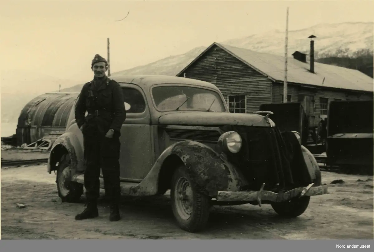 En soldat står fremfor en bil, et hus og en brakke.