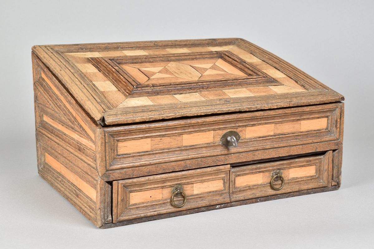 Skrin tillverkat i ek, med rik intarsia av två andra träslag. Rektangulär till formen med pulpetlock. Locket med profilerad och centrerad spegel. Framsida med nyckelhål och nyckelskylt, samt två lådor med draghandtag av mässing. Lådorna går att låsa från skrinets insida med hjälp av två träpluggar. Insidan av skåpet försedd med avsats för placering av skrivredskap samt tre mindre lådor med dragknoppar. 