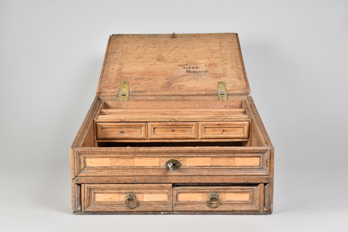 Skrin tillverkat i ek, med rik intarsia av två andra träslag. Rektangulär till formen med pulpetlock. Locket med profilerad och centrerad spegel. Framsida med nyckelhål och nyckelskylt, samt två lådor med draghandtag av mässing. Lådorna går att låsa från skrinets insida med hjälp av två träpluggar. Insidan av skåpet försedd med avsats för placering av skrivredskap samt tre mindre lådor med dragknoppar. 